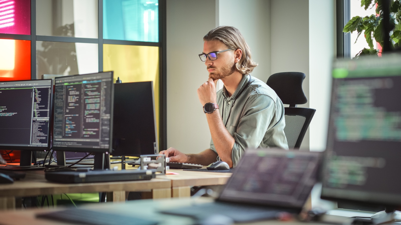 An engineer in glasses, hand on chin, reviewing work on his monitors in a bright office